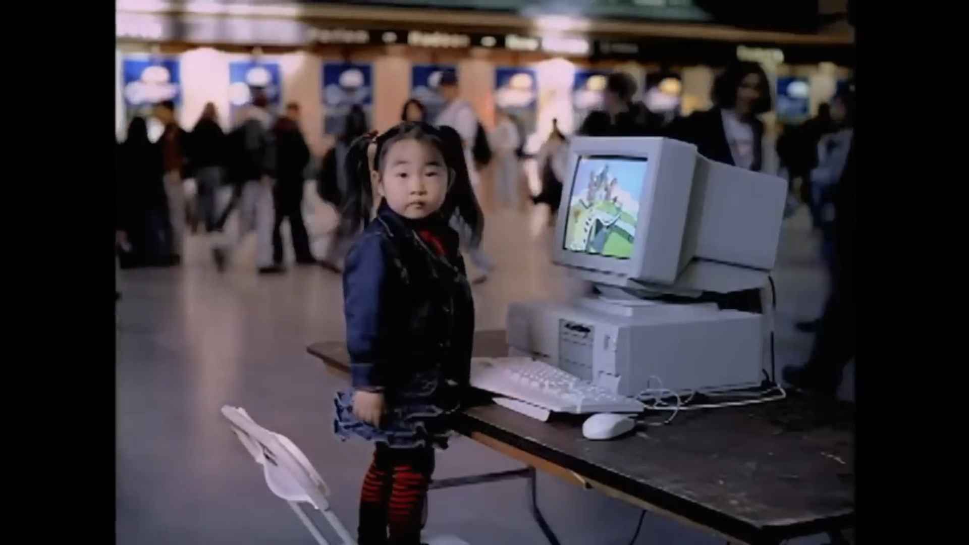 A little girl stood next to a computer running Windows 95, in an apparent tube station